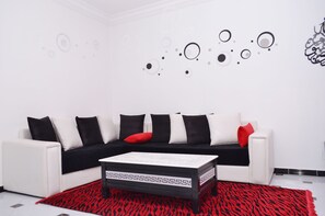 A modern living room with a white sofa, black and red cushions, a patterned coffee table, and a red and black rug. - Chic T3 Akid Lotfi WIFI (Bir El Djir)