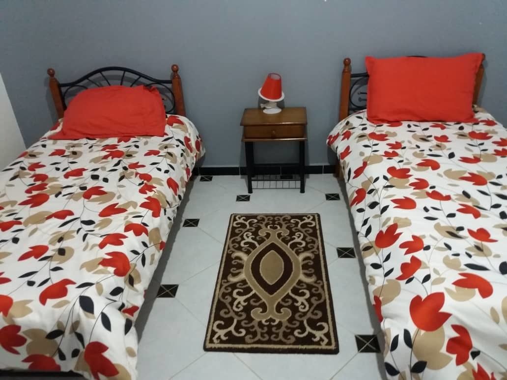 A room with two beds, each with a floral bedspread and a red pillow. There is a small wooden nightstand with a red lamp between the beds. A decorative rug is placed in the center of the room.