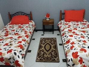 A room with two beds, each with a floral bedspread and a red pillow. There is a small wooden nightstand with a red lamp between the beds. A decorative rug is placed in the center of the room. - Chic T3 Akid Lotfi WIFI (Bir El Djir)