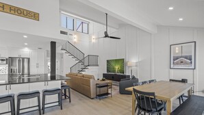 Et rummeligt køkken med moderne hårde hvidevarer, et spiseområde med et træbord og stole, en hyggelig stue med sofa og en stor, grøn plante, samt en trappe til en etage ovenpå.