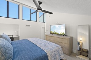 Ein Schlafzimmer mit Bett, Fernseher, Kommode, Deckenventilator und großen Fenstern.