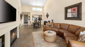 Uma sala de estar ampla com lareira, um sofá de couro, uma área de refeições com um lustre e uma cozinha ao fundo.