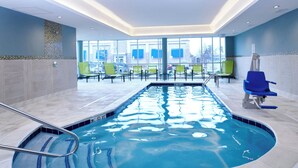 Ein Innenpool mit freiem Blick ins Freie durch große Fenster, ein gefliestes Becken und mehrere grüne Stühle rund um den Pool.