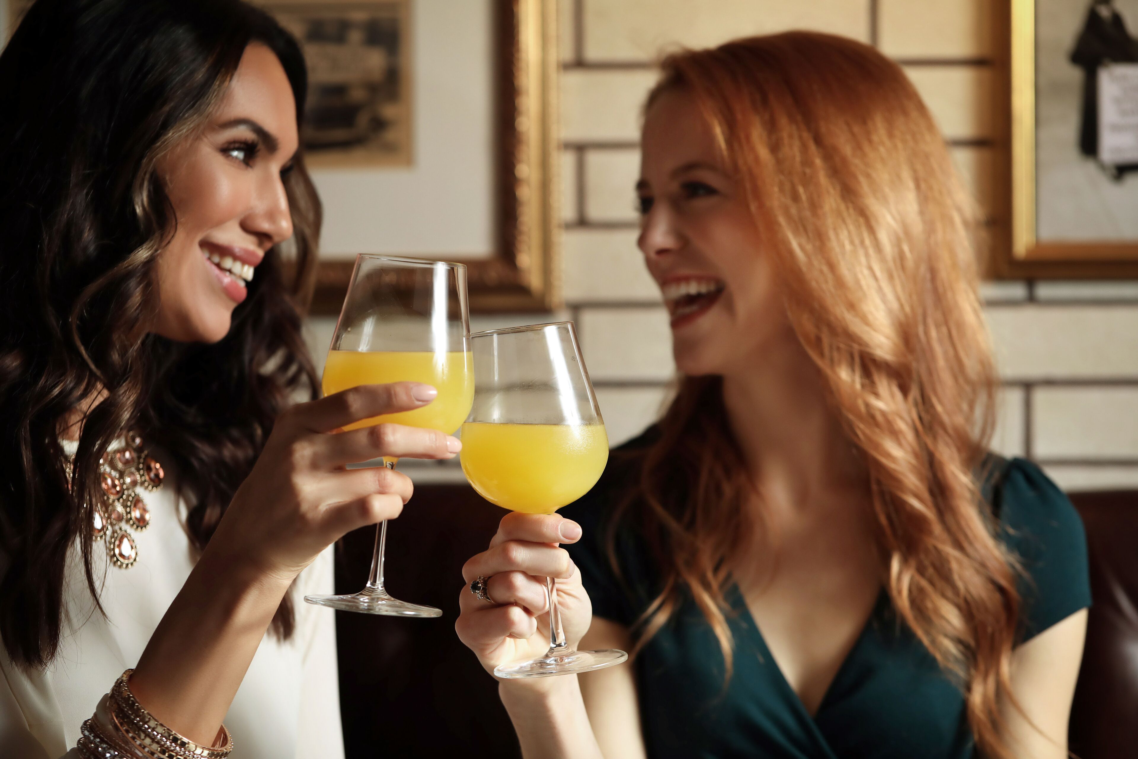 Two women toasting with glasses of orange juice in a room with a brick wall and framed pictures.