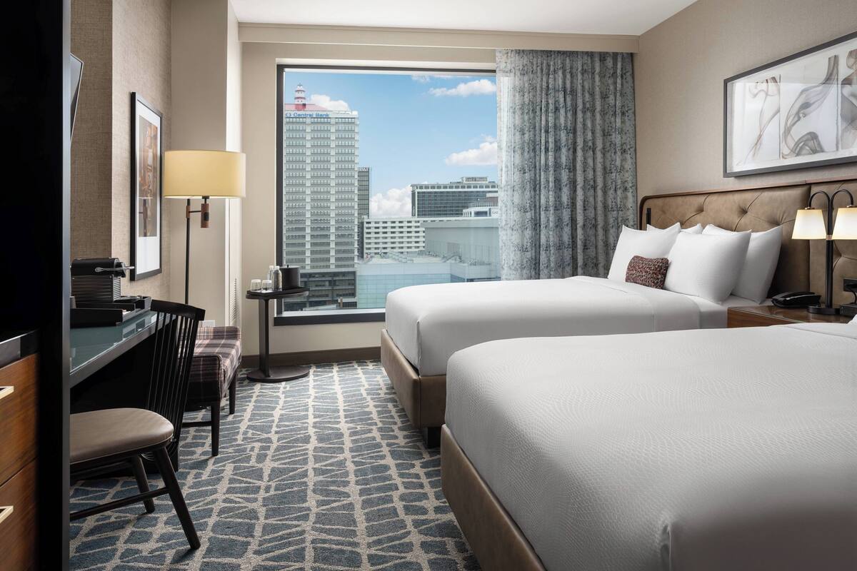 A hotel room with two beds, a desk, a chair, a window with a city view, and a patterned carpet.