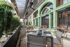 An outdoor dining area with tables and chairs, a green building facade, and a row of plants along the wall.