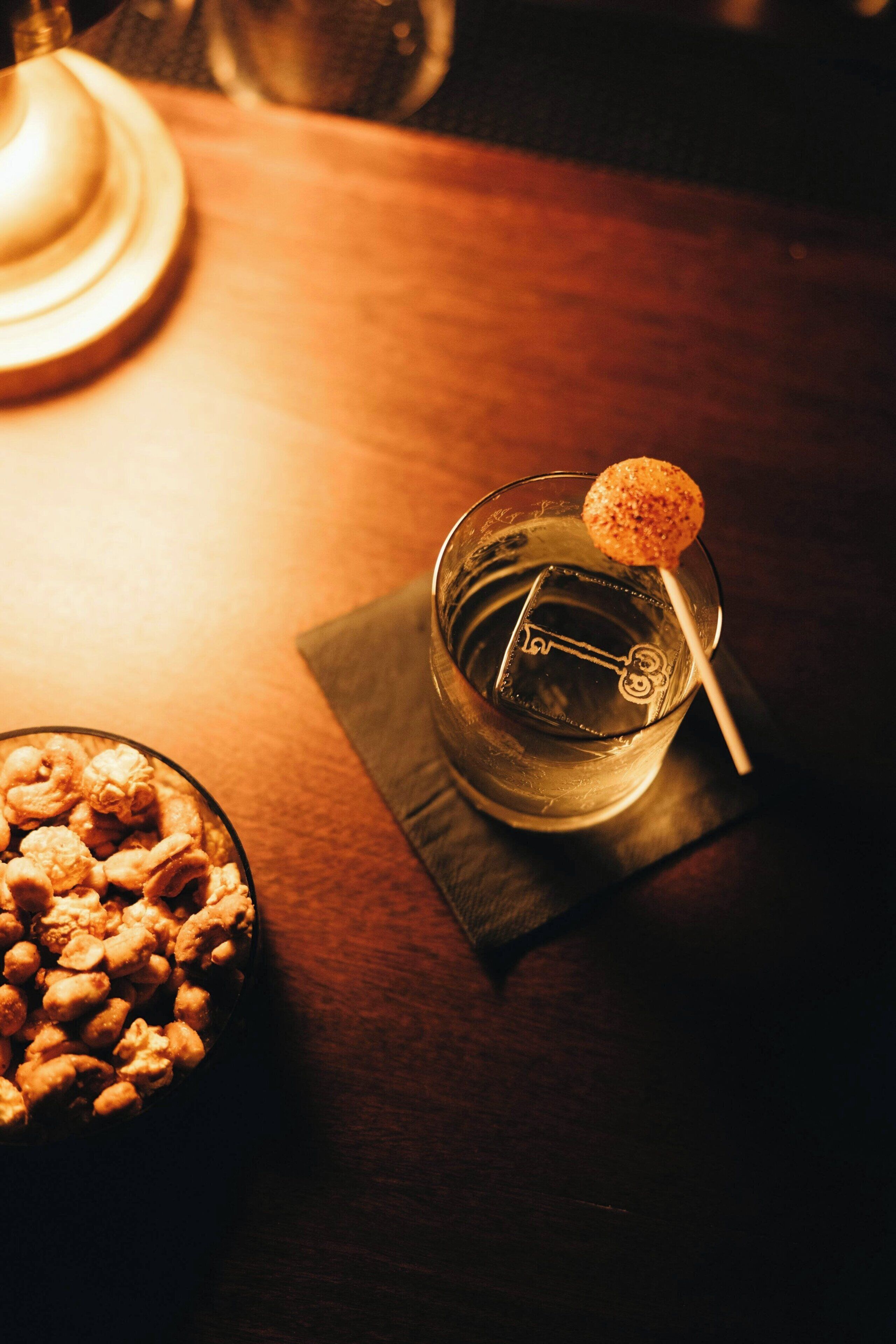 A glass with a cocktail, a key-shaped ice cube, and a lollipop on a napkin on a wooden table.