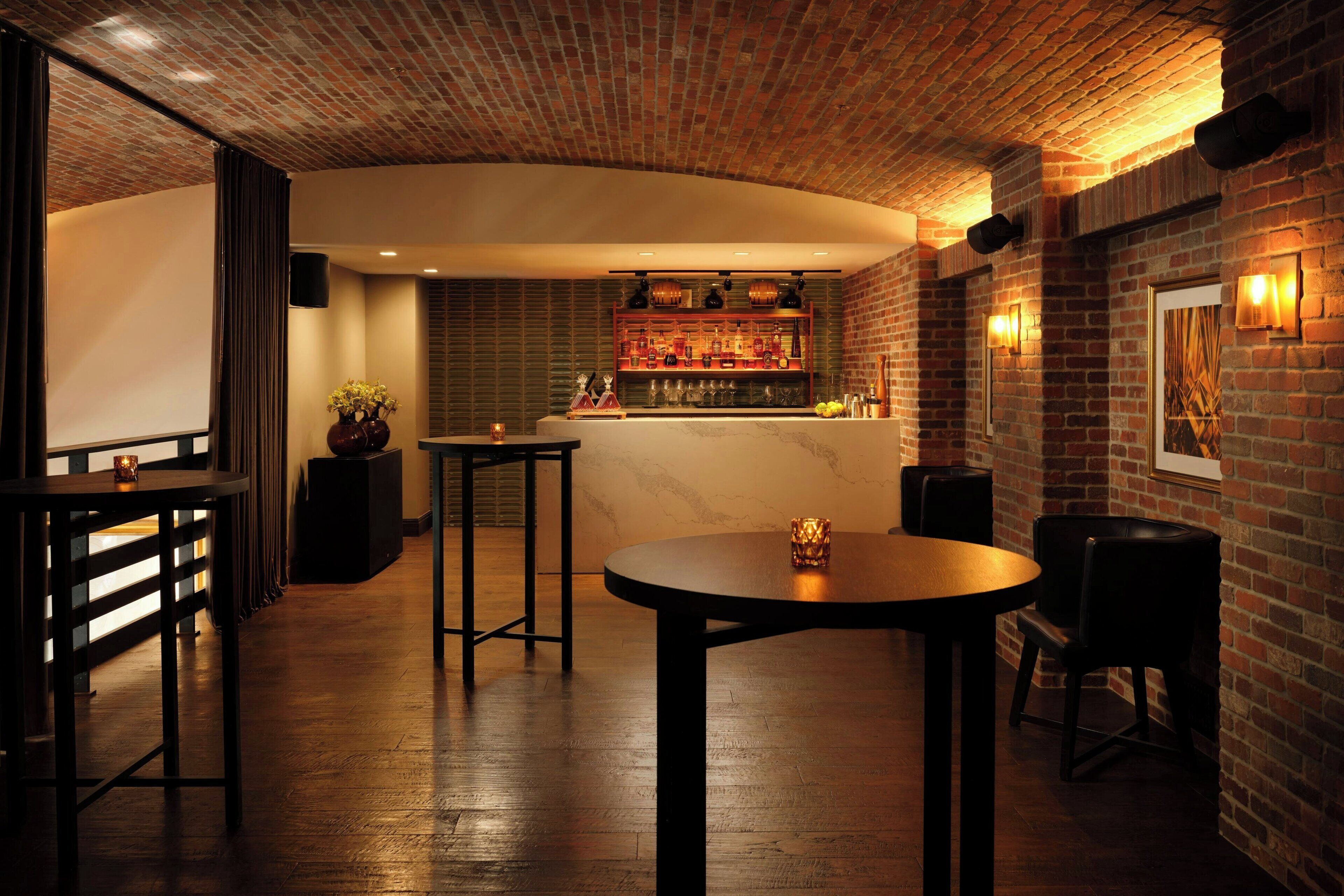 Uma área de bar com teto de tijolos, piso de madeira e balcão com copos de vidro. Há mesas e cadeiras para acomodação.