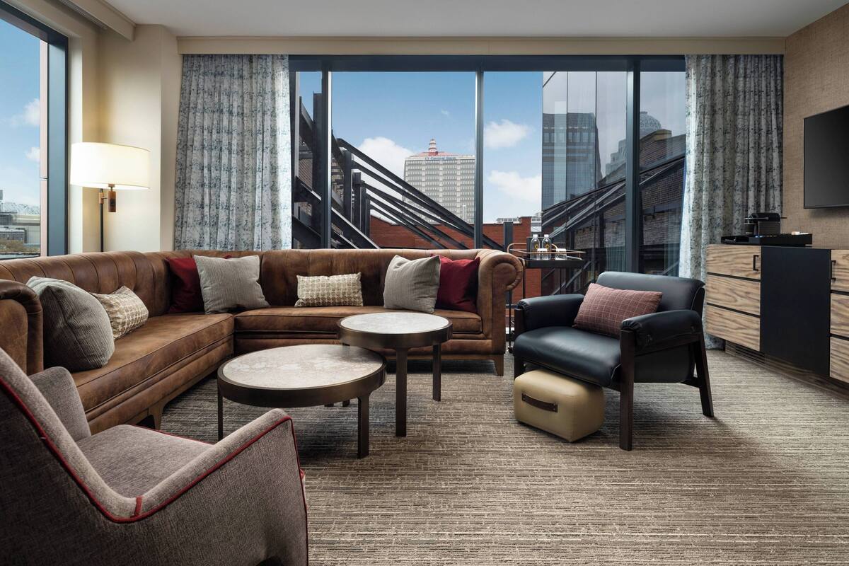 A modern hotel room with a sectional sofa, a leather armchair, a round coffee table, and a TV mounted on the wall.