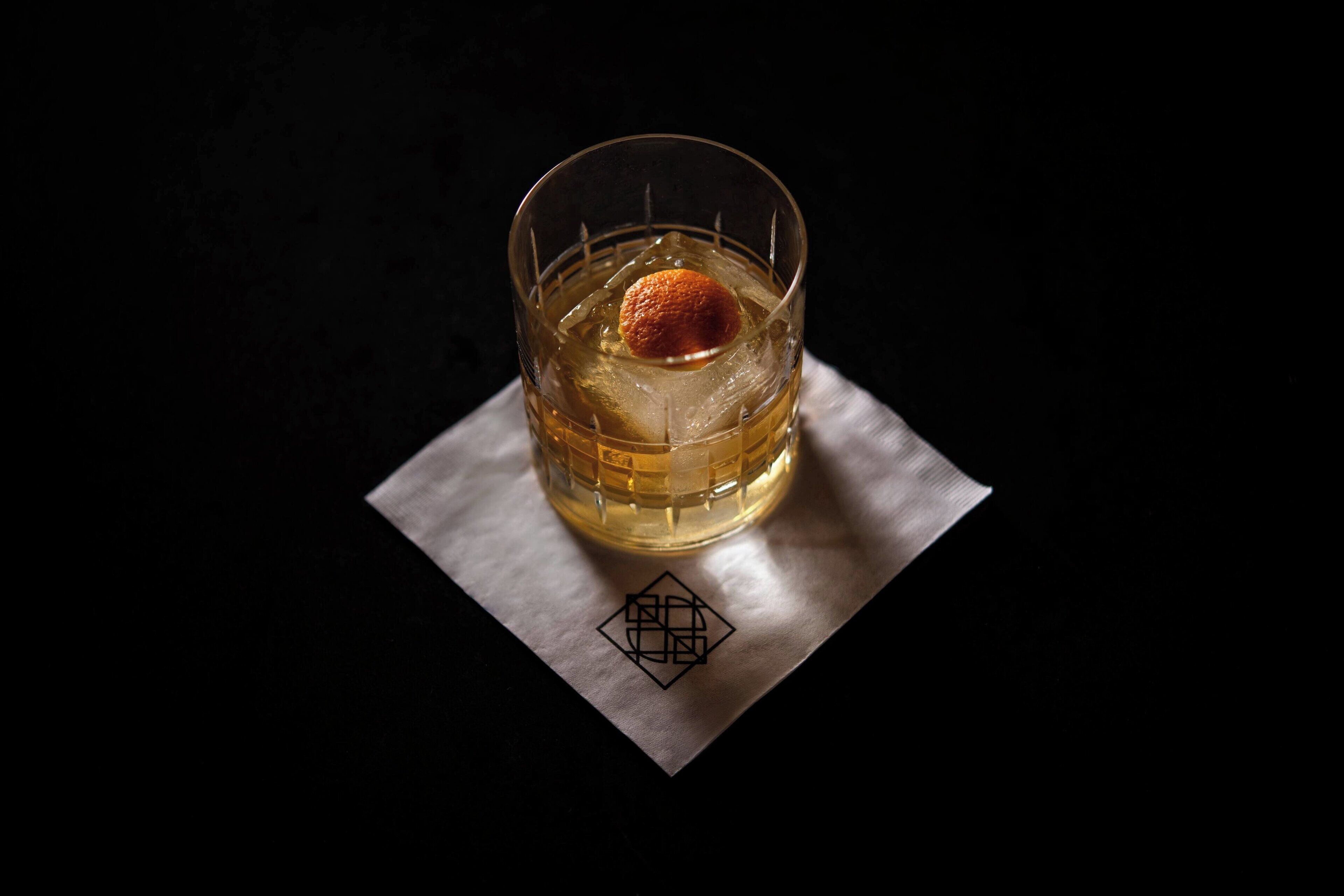 A glass of amber liquid with a spherical garnish on a coaster.