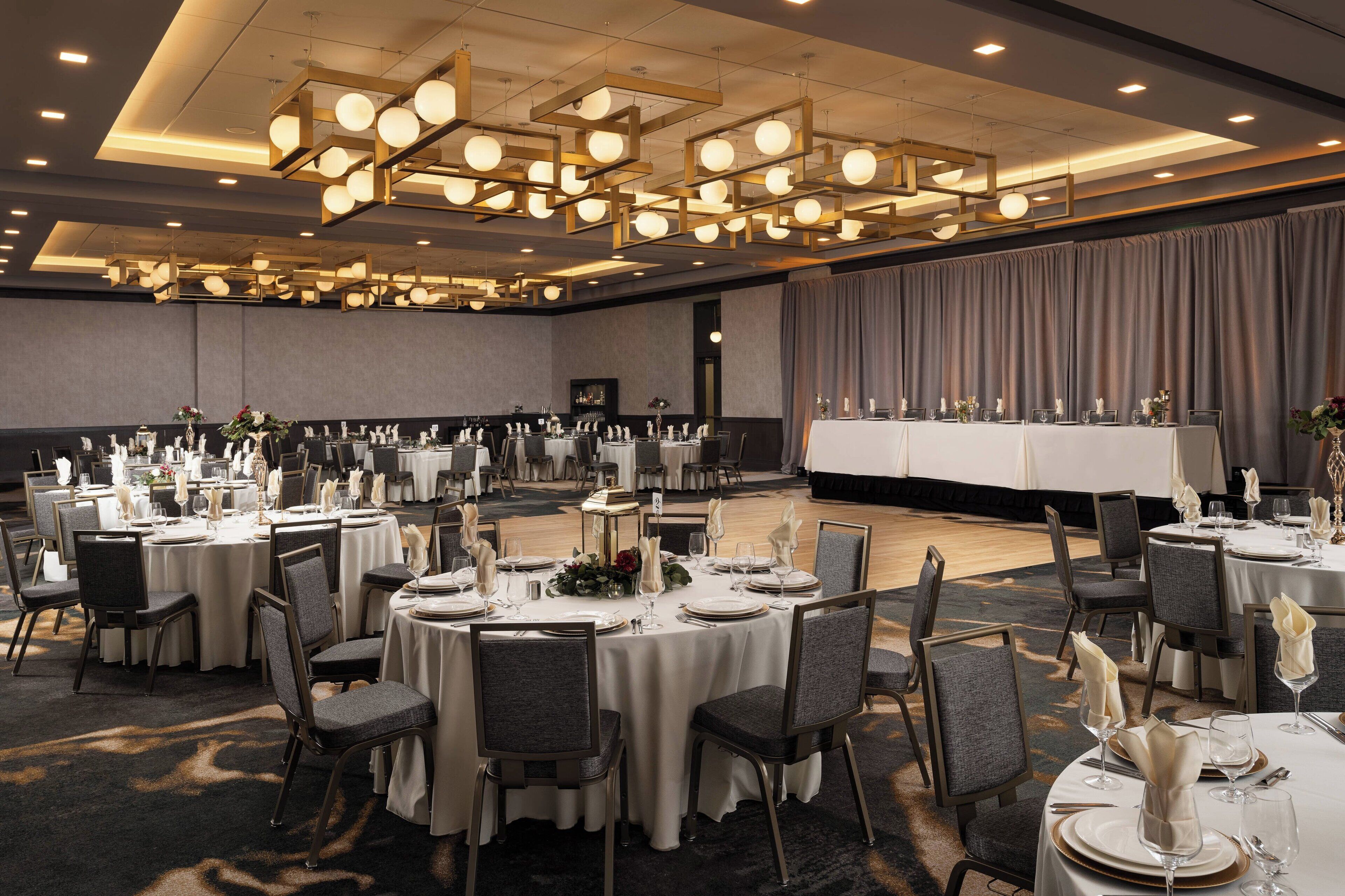 A large banquet hall with round tables set for dining, featuring chandeliers and a stage area.