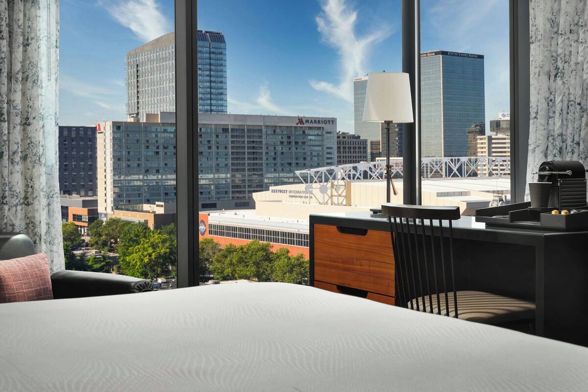 A hotel room with a large window offering a city view, a bed, a desk, and a chair.