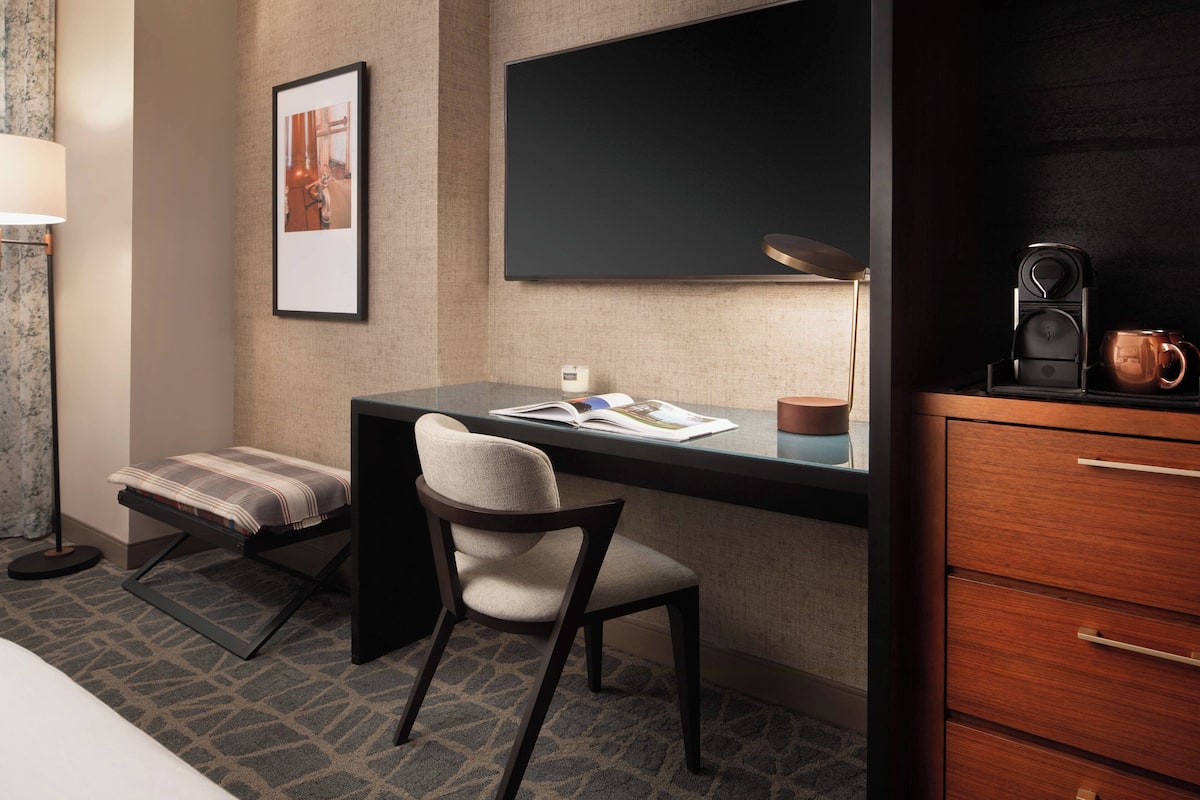 Quarto de hotel com escrivaninha, cadeira e máquina de café em uma mesinha lateral.
