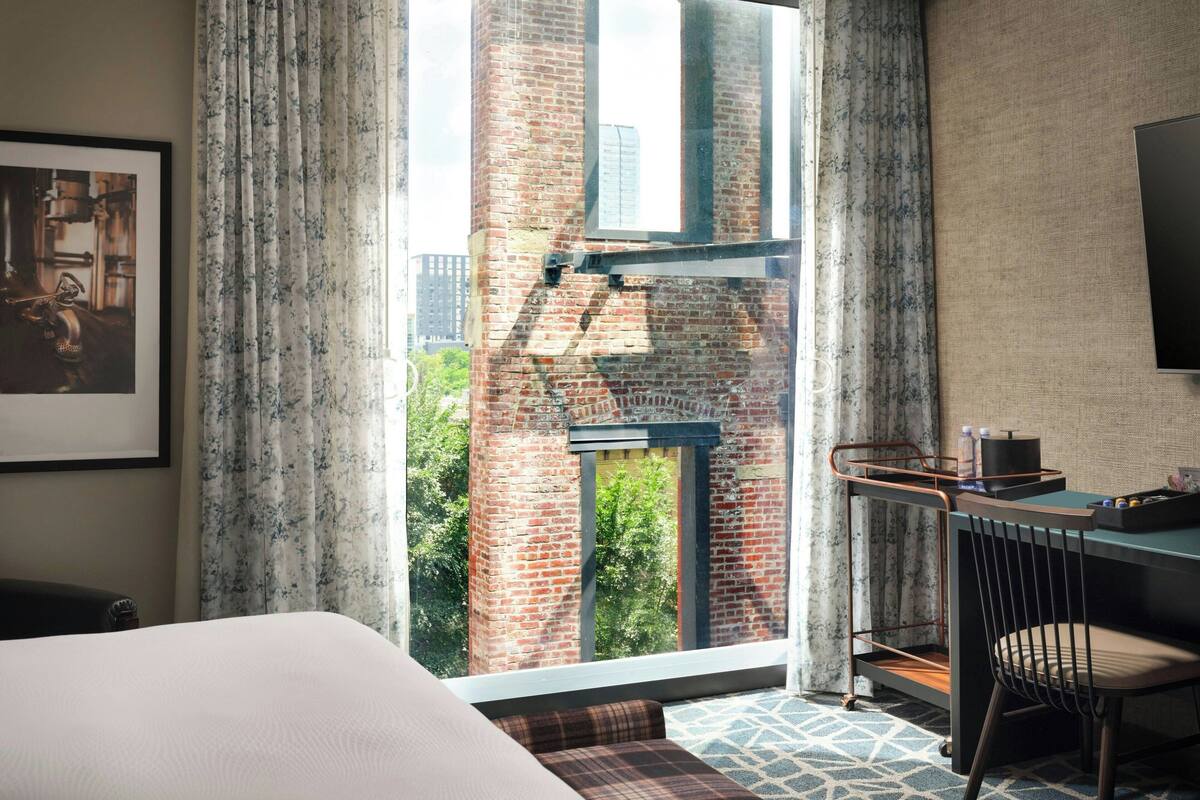 A hotel room with a large window, a bed, a desk with a chair, and a mounted television.