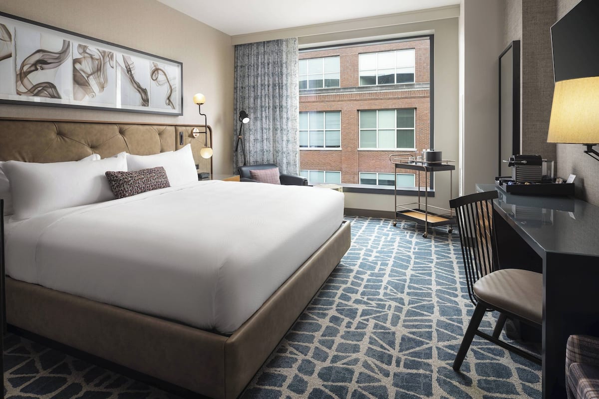 A hotel room with a large bed, a desk, a chair, and a view of a brick building through the window.