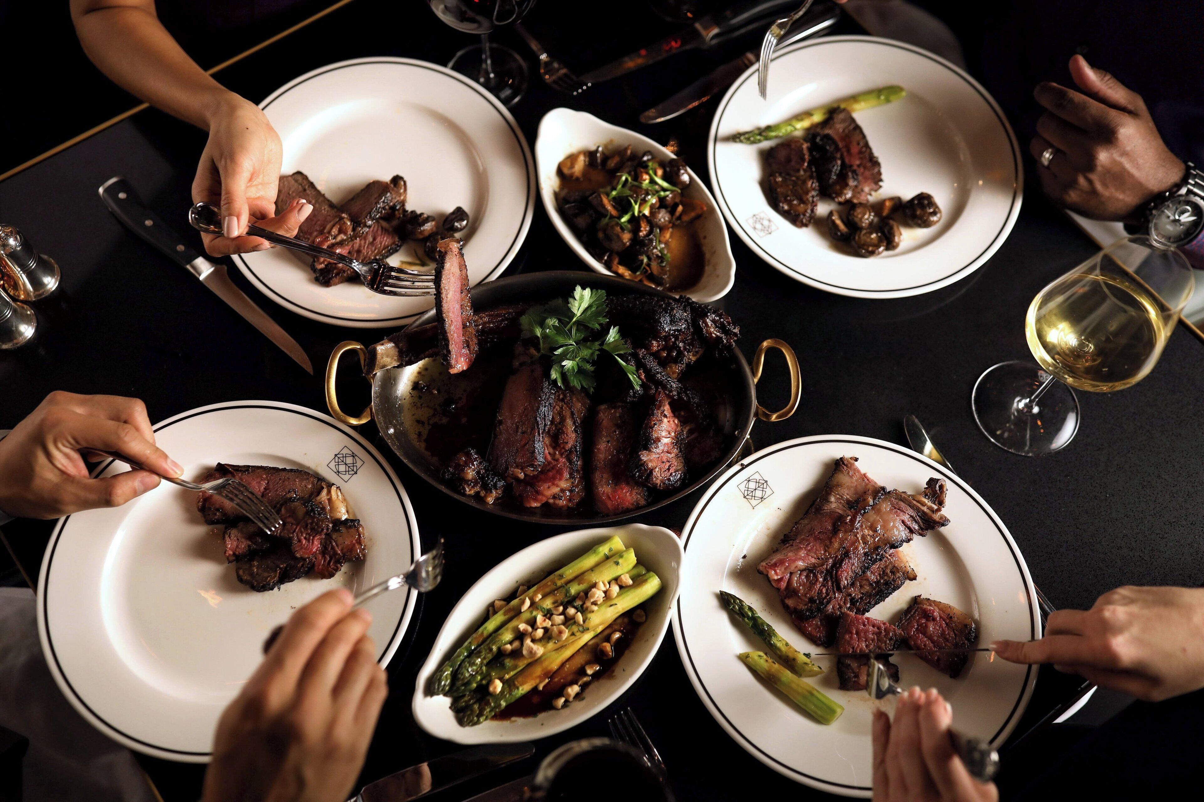 A group of people dining around a table with multiple plates of food, including steak and asparagus.