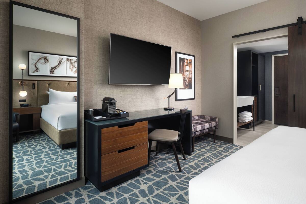 A hotel room with a bed, a desk with a chair, a TV mounted on the wall, and a large mirror.