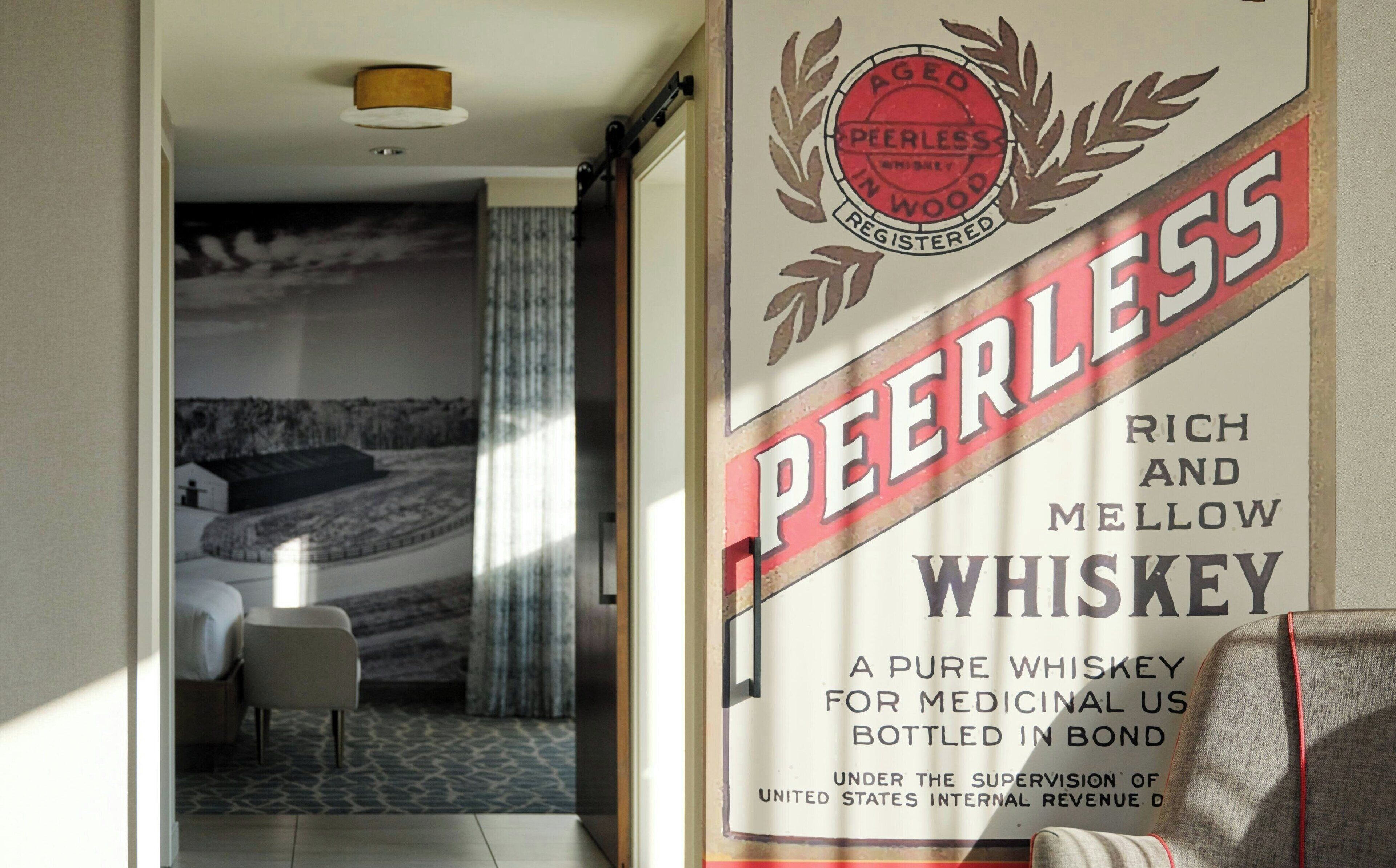 Una habitación de hotel con una cama, una silla y una ventana. Un cartel en la pared anuncia el whiskey Peerless.
