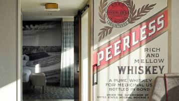 Una habitación de hotel con una cama, una silla y una ventana. Un cartel en la pared anuncia el whiskey Peerless.