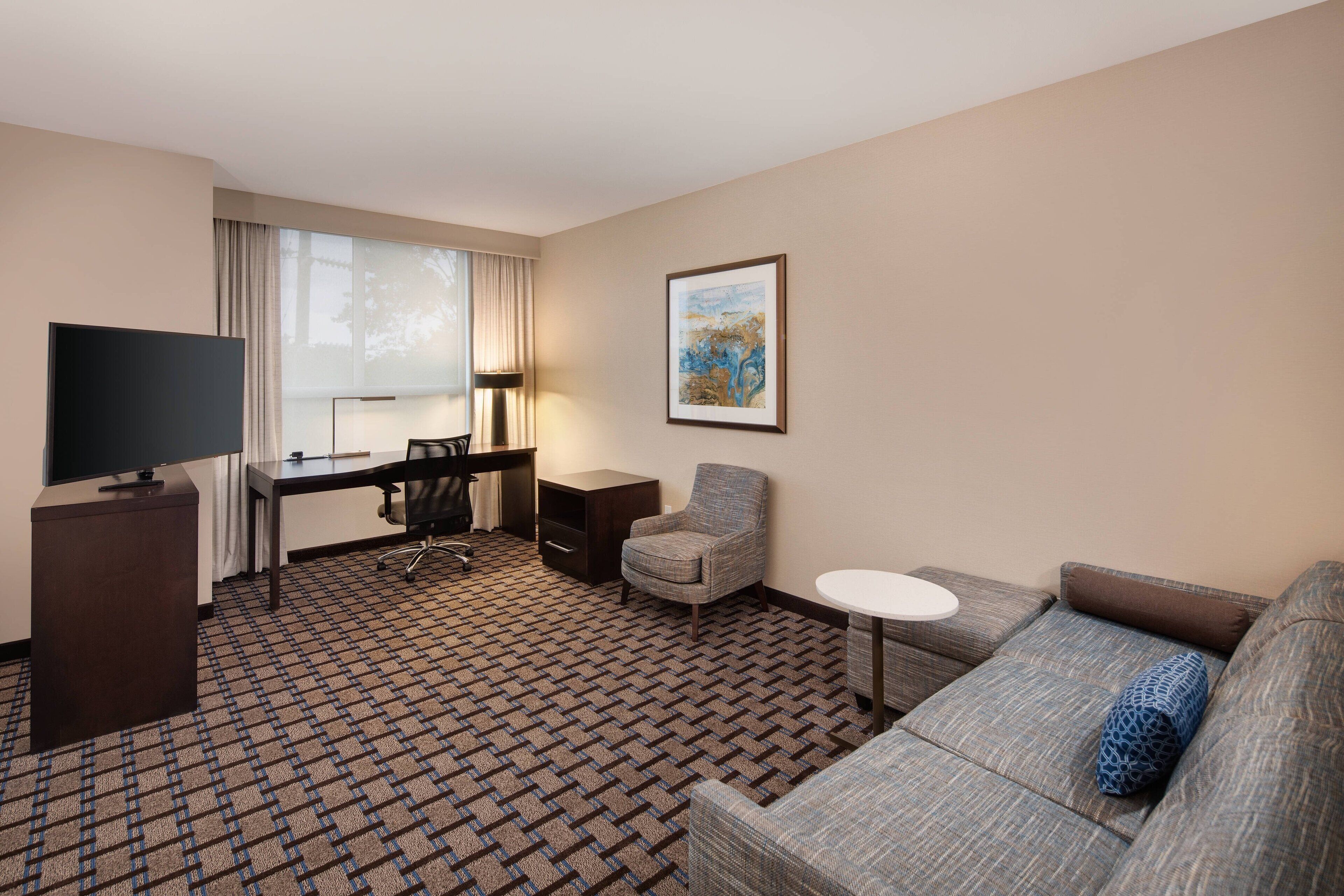 A hotel room with a large flat-screen TV, a desk with a chair, a sofa, and a small round table.