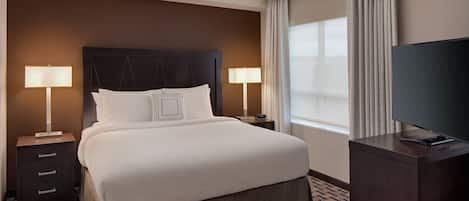 Quarto de hotel com uma cama grande, duas mesas de cabeceira com luminárias, uma TV de tela plana em um suporte e uma janela com cortinas.