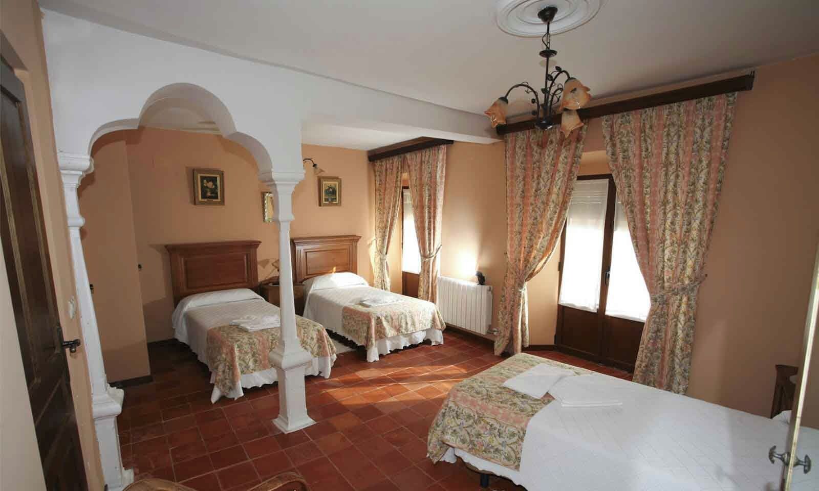 Une chambre d’hôtel avec deux lits, une grande fenêtre avec des rideaux, un radiateur et un lustre.