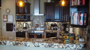 A kitchen with dark cabinets, a granite countertop, a stove, and a coffee maker. There are two bar stools and a shelf with books. - Downtown Penthouse-Style Apartment (Puerto Vallarta)
