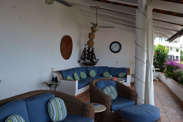 Ein überdachter Außenbereich mit blauen, gepolsterten Sofas und Sesseln, einer an der Wand befestigten Uhr und einem dekorativen Schiffmodell.