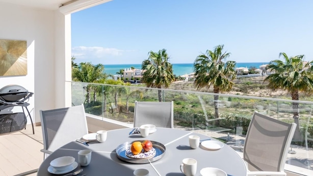 A balcony with a table set for breakfast, overlooking a coastal view with palm trees and houses.