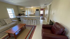 A living room with a sofa, a coffee table, and a red leather couch. There is a kitchen area with a refrigerator, microwave, and bar stools.