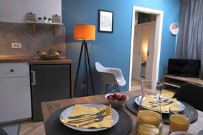 Una cocina moderna con comedor, un refrigerador pequeño, una cesta de fruta y una mesa preparada para el desayuno, con platos, cubiertos y vasos de jugo de naranja.