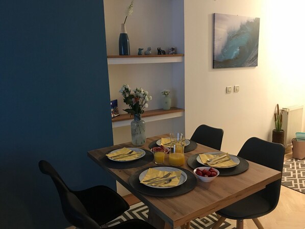 Comedor con una mesa de madera dispuesta para cuatro personas, rodeada de sillas negras. Hay platos, cubiertos y un frutero sobre la mesa. Un jarrón con flores está en la pared encima de la mesa.