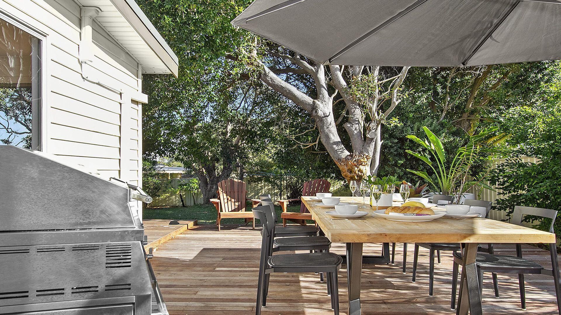 An outdoor patio with a wooden table and chairs, surrounded by greenery and a large tree.