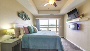 Un dormitorio con cama, mesita de noche, ventilador de techo, televisor en un estante y vista al exterior.