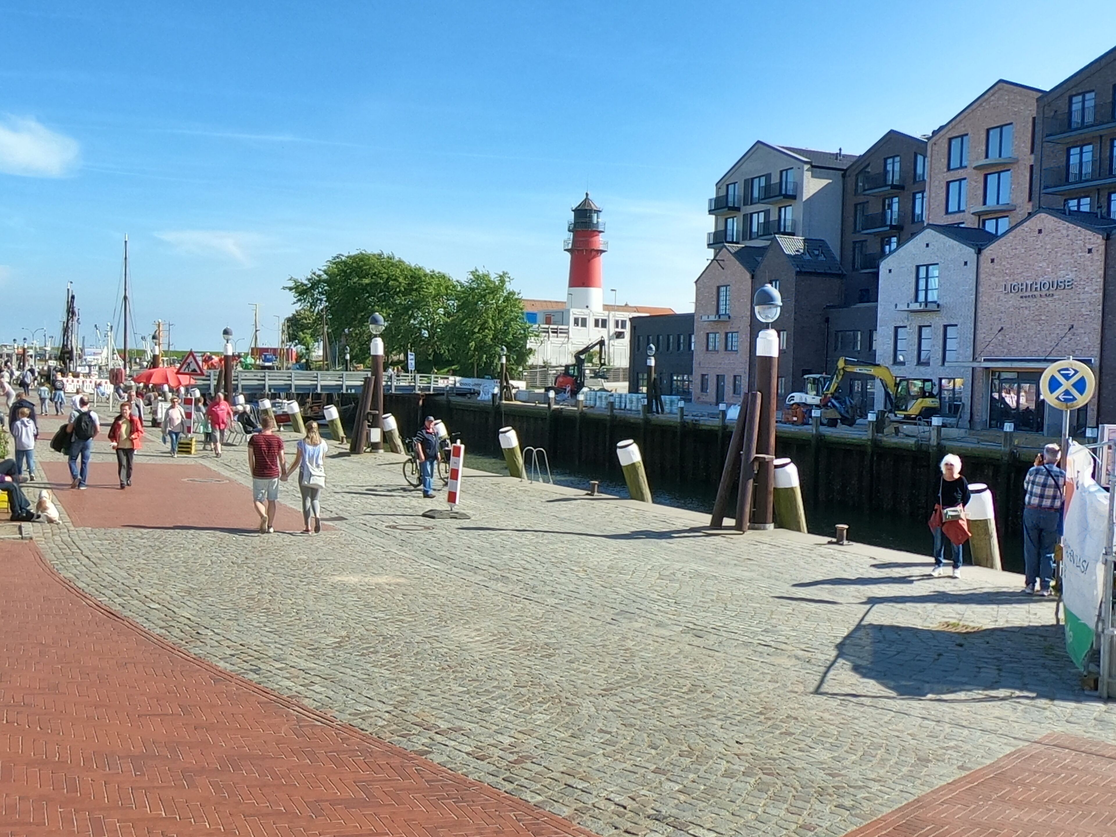 A waterfront promenade with a red brick pathway, a lighthouse, and modern buildings with balconies.