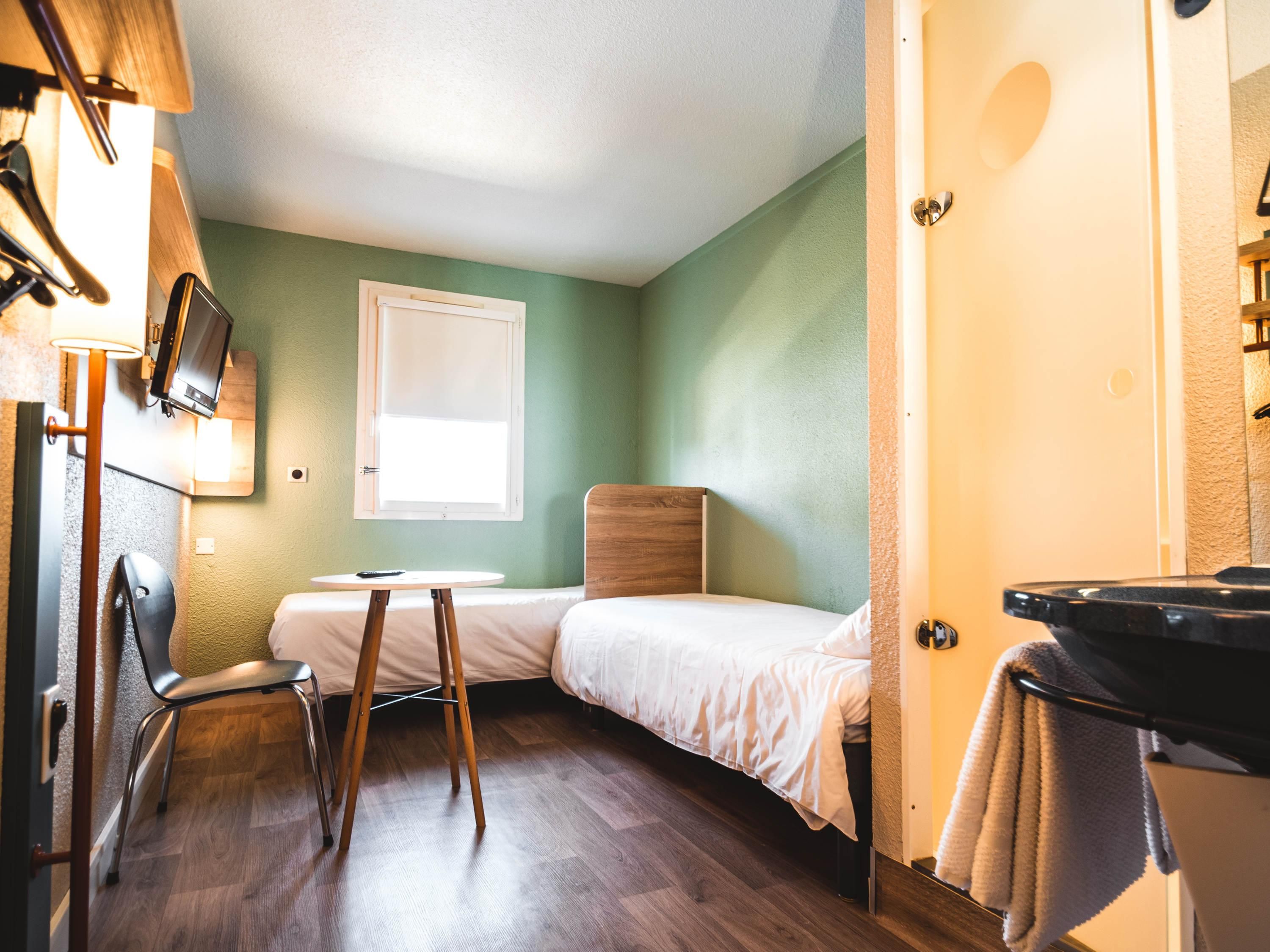 Una habitación pequeña con cama individual, cabecera de madera, una mesita, una silla, televisor montado en la pared y una ventana.
