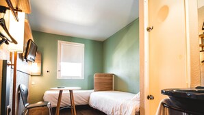 A small, single-bed room with a wooden headboard, a small table, a chair, a TV mounted on the wall, and a window. - Ibis Budget Saint Brieuc Yffiniac (Yffiniac)