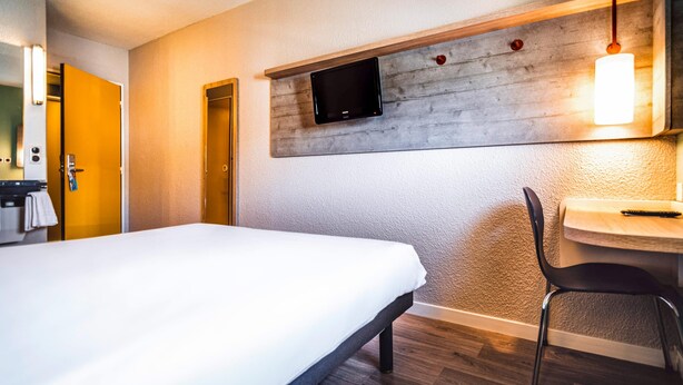 A hotel room with a bed, a desk, a chair, a television mounted on the wall, and a door. - Ibis Budget Saint Brieuc Yffiniac (Yffiniac)
