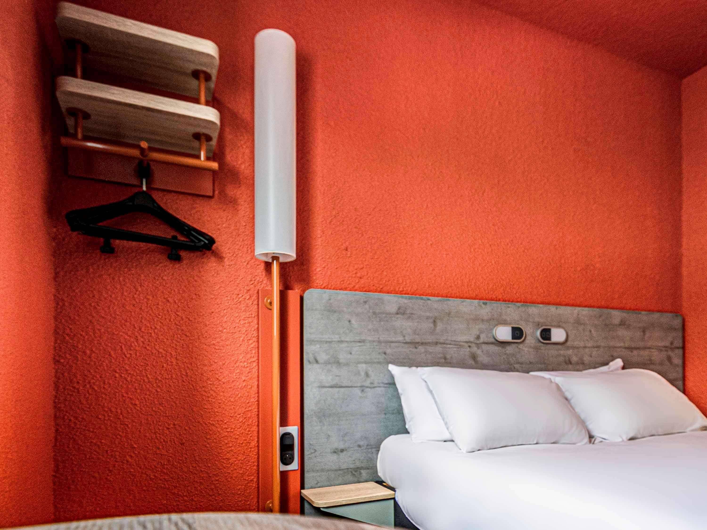 Une chambre d’hôtel avec un mur rouge à texture, une tête de lit en bois, un lit avec une literie blanche et un objet cylindrique blanc accroché au mur.