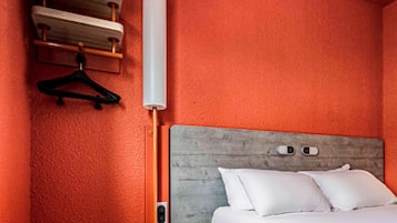 Une chambre d’hôtel avec un mur rouge à texture, une tête de lit en bois, un lit avec une literie blanche et un objet cylindrique blanc accroché au mur.