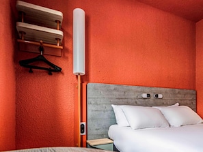 A hotel room with a red textured wall, a wooden headboard, a bed with white bedding, and a white cylindrical object on the wall. - Ibis Budget Saint Brieuc Yffiniac (Yffiniac)