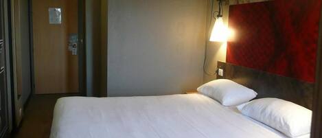 A hotel room with a bed, a wall-mounted lamp, and a door with a sign.