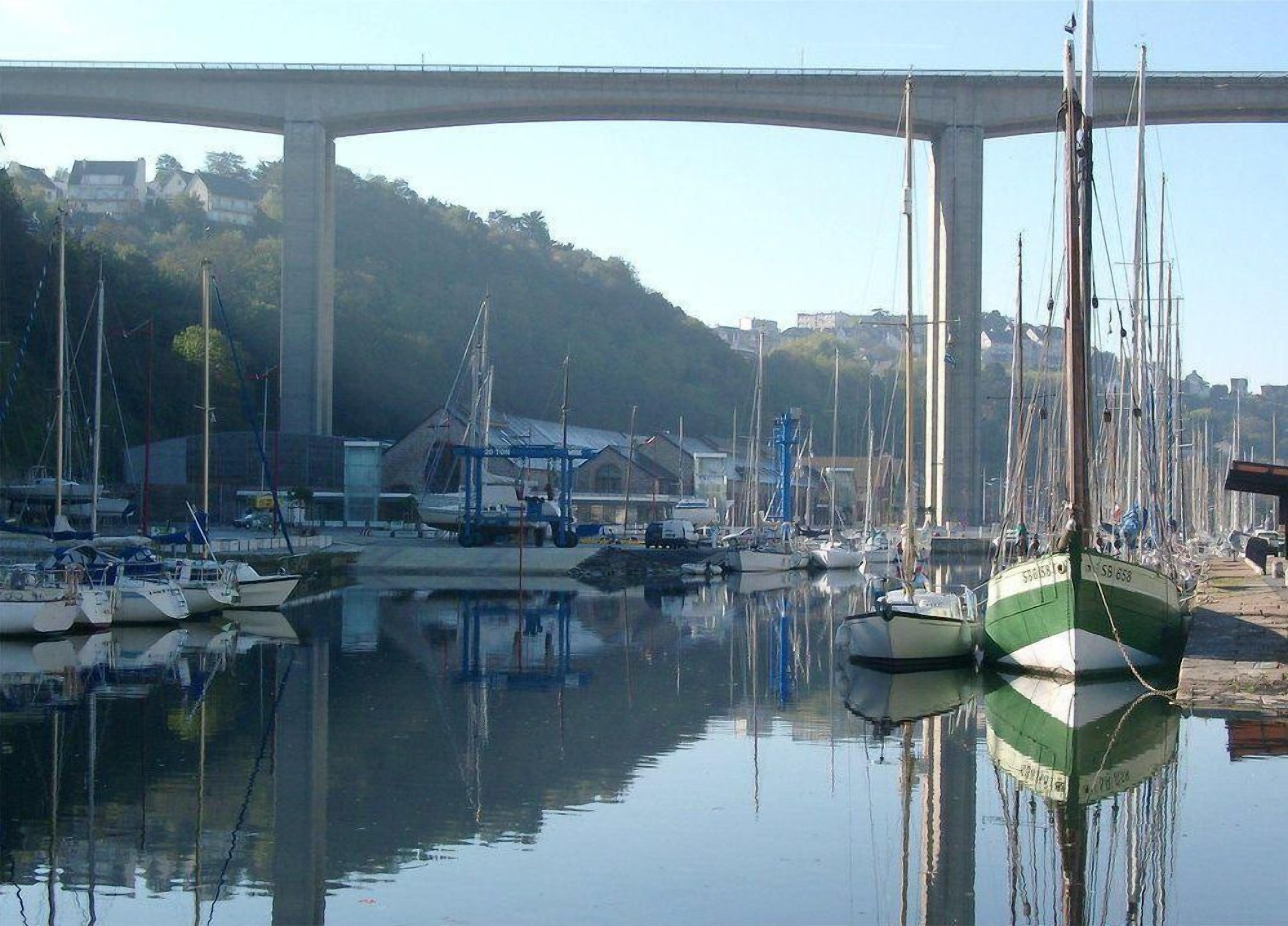 Un puerto deportivo con barcos amarrados, un puente al fondo y un paisaje montañoso.
