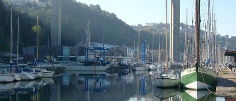 A marina with boats docked, a bridge in the background, and a hilly landscape.