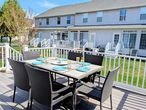 A deck with a table set for four, surrounded by wicker chairs, overlooking a house with a white fence.