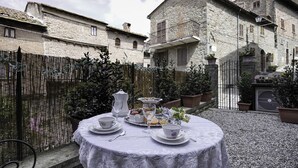 Una mesa redonda dispuesta para una comida, con platos, tazas y una tetera, rodeada de plantas en macetas y un edificio de piedra al fondo.