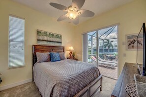 A bedroom with a bed, a ceiling fan, a window with blinds, a painting, and a view of the outdoors.