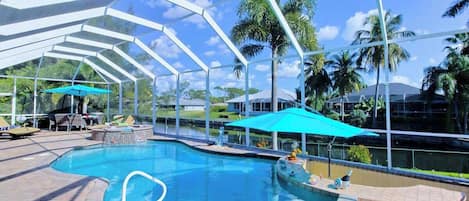 A covered outdoor swimming pool with a clear sky, palm trees, and a bar area.