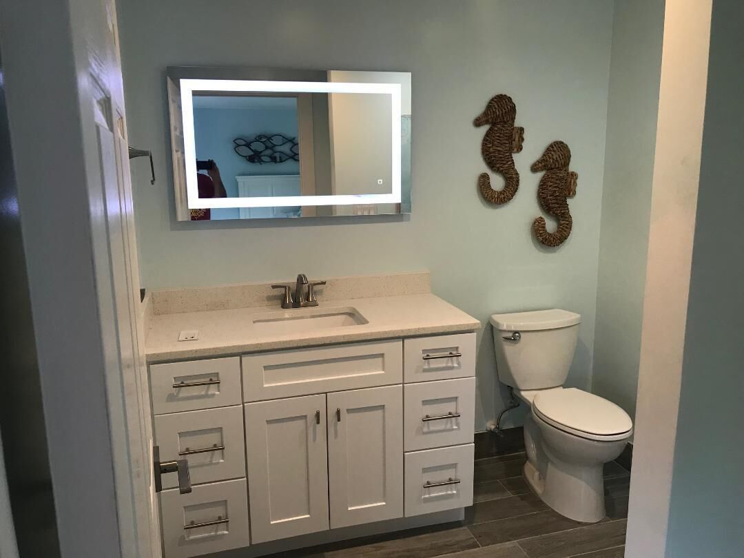 A bathroom with a white vanity, a mirror, and a toilet. There are decorative wall hangings above the toilet.
