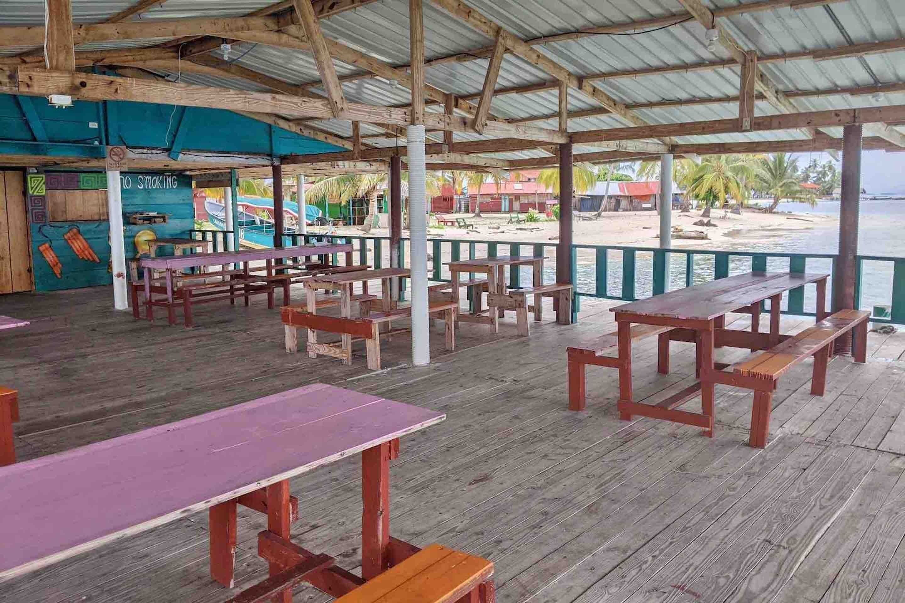Un área de comedor al aire libre con mesas y bancos de madera, techo cubierto y vista a la playa y al océano.